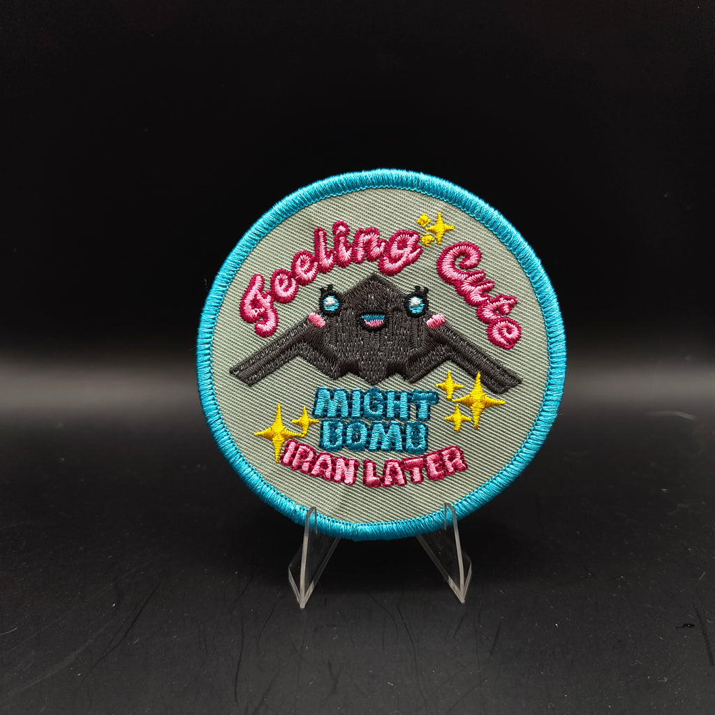 Embroidered patch with text and a B2 Bomber Kawaii Cute "Feeling Cute Might bomb Iran later" Text design on a stand against a black background