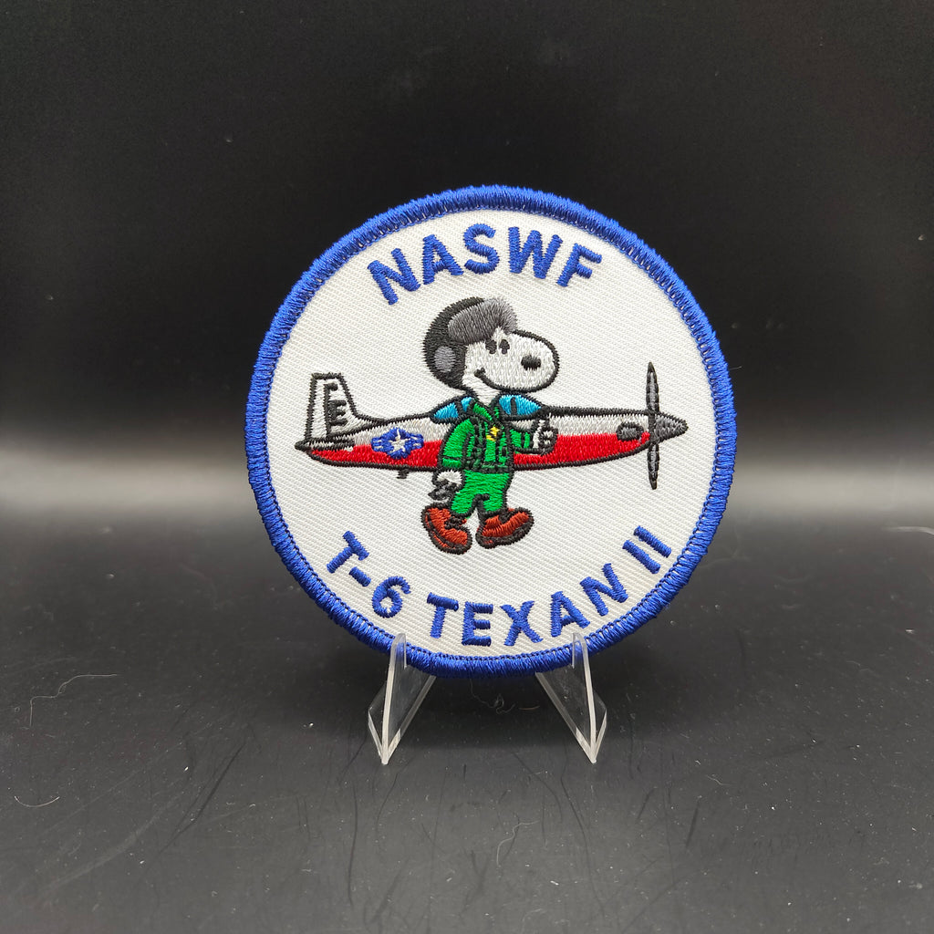 Patch featuring a cartoon character holding an airplane with 'NASWF T-6 TEXAN II' text on a black background Snoopy dog peanuts